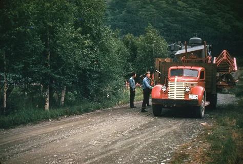 Men standing by parked red truck on gravel road.